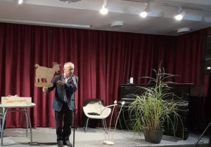 Pan Eugeniusz Dolat stoi na scenie recytując wiersz. Autor trzyma w ręku kukiełkę kozy - postać z prezentowanego utworu.